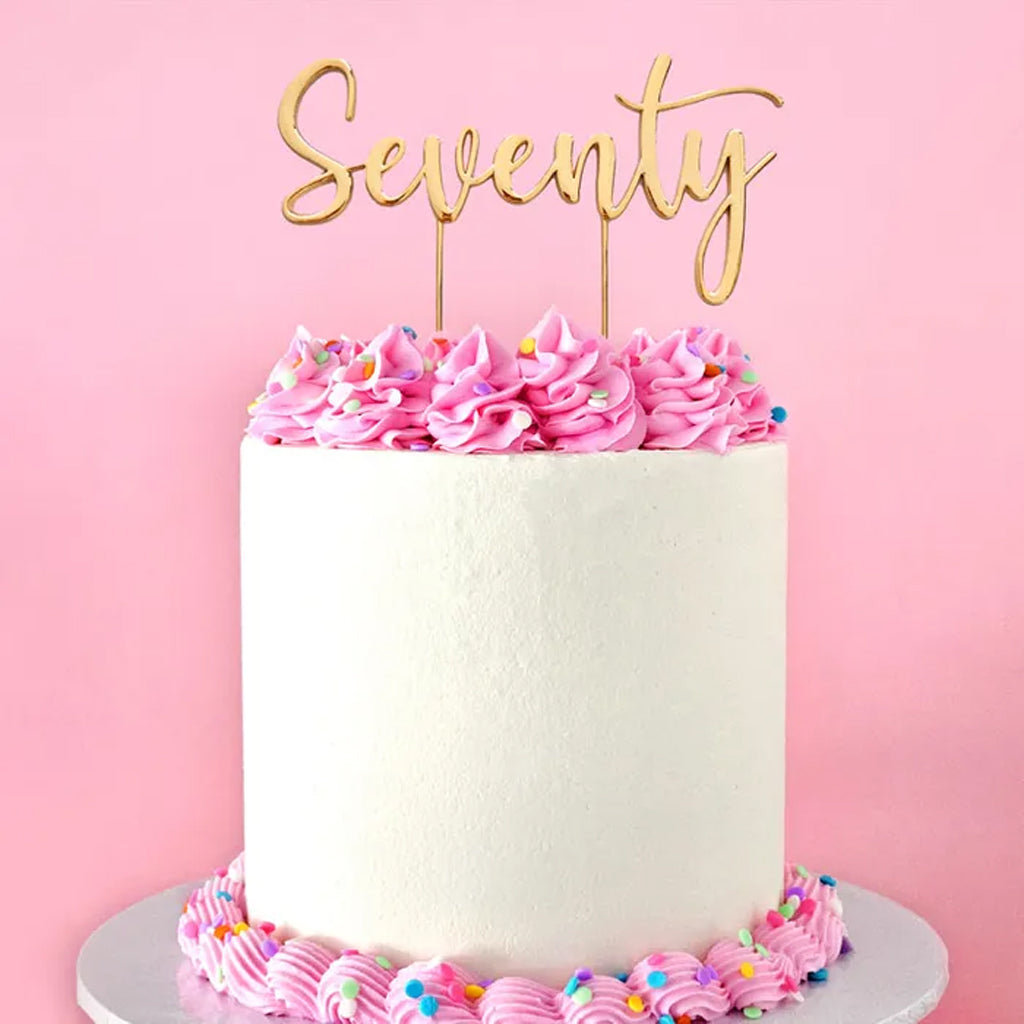 A white frosted birthday cake with pink piped frosting rosettes and sprinkles, topped with a gold 'Seventy' script cake topper, set against a solid pink background.