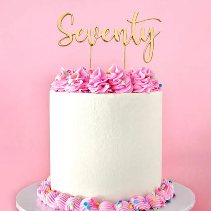 A white frosted birthday cake with pink piped frosting rosettes and sprinkles, topped with a gold 'Seventy' script cake topper, set against a solid pink background.