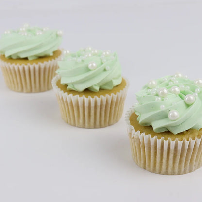 Three mini cupcakes with light mint green frosting, white pearl sprinkles, and white paper liners, arranged on a light gray background.