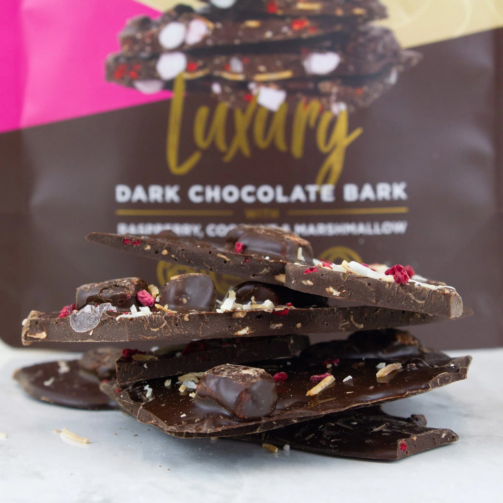A stack of dark chocolate bark pieces with raspberry, coconut, and marshmallow inclusions, placed in front of a branded bag labeled 'Luxury Dark Chocolate Bark Raspberry Coconut & Marshmallow'.