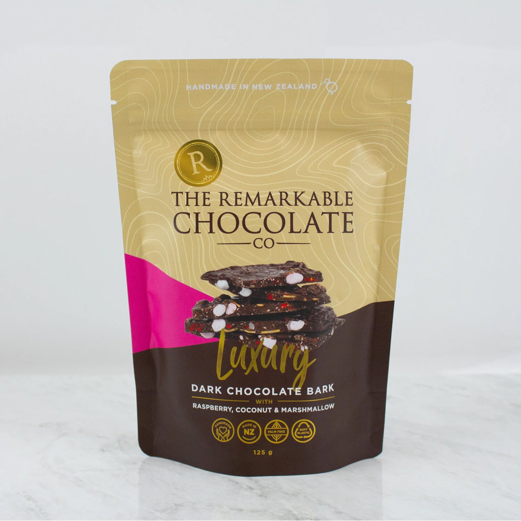 A stand-up pouch of The Remarkable Chocolate Co. Luxury Dark Chocolate Bark, featuring a gold and brown design with a pink accent and an image of chocolate bark with marshmallows, coconut and raspberry pieces, set against a light gray background.