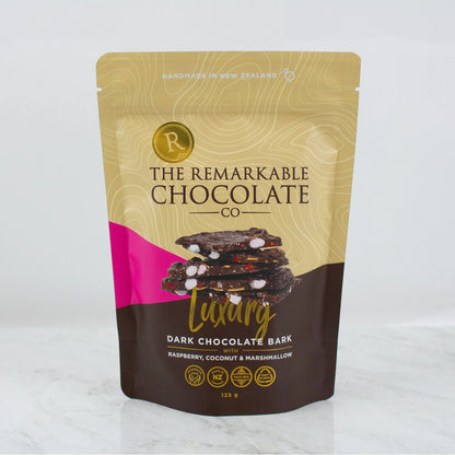 A stand-up pouch of The Remarkable Chocolate Co. Luxury Dark Chocolate Bark, featuring a gold and brown design with a pink accent and an image of chocolate bark with marshmallows, coconut and raspberry pieces, set against a light gray background.