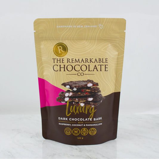A stand-up pouch of The Remarkable Chocolate Co. Luxury Dark Chocolate Bark, featuring a gold and brown design with a pink accent and an image of chocolate bark with marshmallows, coconut and raspberry pieces, set against a light gray background.