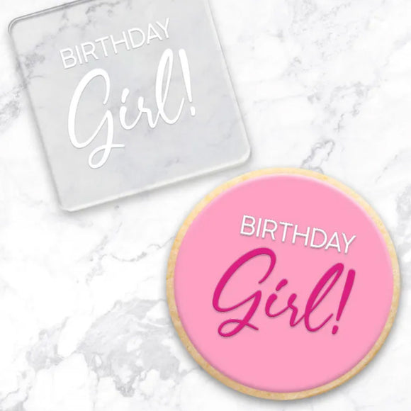 clear debosser with a Birthday Girl! pattern beside a cookie with pink icing and pink and white writing on a marble background