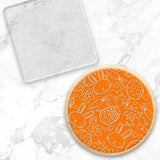 clear debosser with a bunny & egg pattern beside a cookie with orange icing and white bunny egg pattern on a marble background
