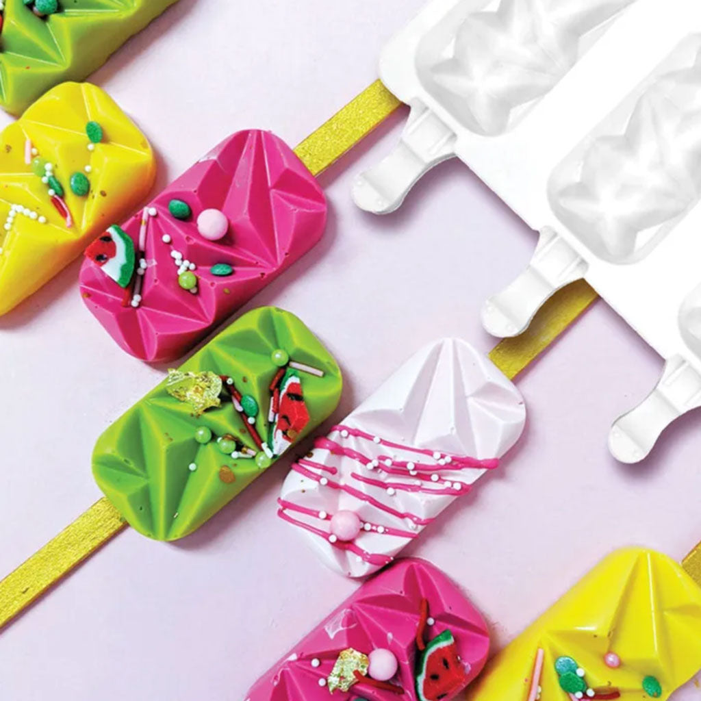 green, pink, yellow and white geo cake pops covered in pink, green and white sprinkles with gold acrylic popsicle sticks lined up beside white silicone mould on a pink background