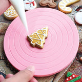 Christmas tree cookie being decorated with white icing on mini pink cookie turntable with other Christmas cookies surrounding 