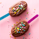 two brown cake pops with pastel coloured sprinkles scattered and pink and blue glitter popsicle sticks on pink background