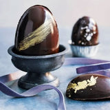 smooth 50g Easter eggs made from dark chocolate with a streak of gold paint in a grey egg cup beside a half egg decorated with gold leaf and purple ribbon
