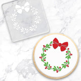 clear debosser with a Christmas wreath pattern beside a cookie with white icing and a green and red Christmas wreath on a marble background