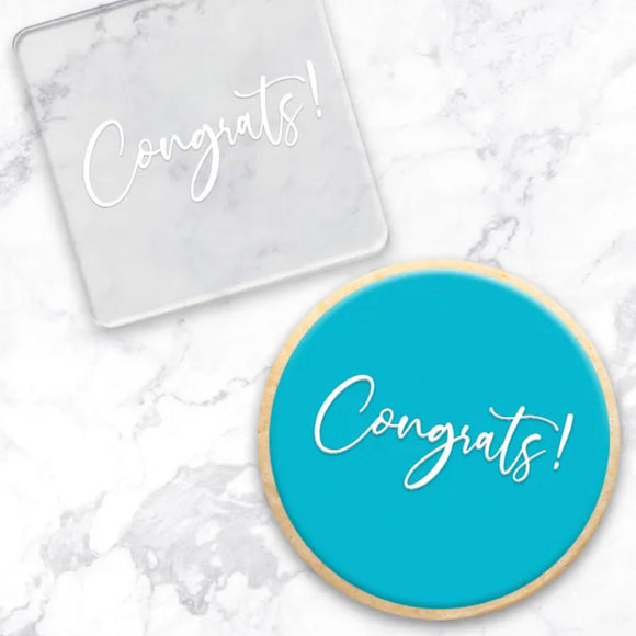 clear debosser with congrats! writing pattern beside a cookie with blue icing and white congrats! written displayed on a marble background