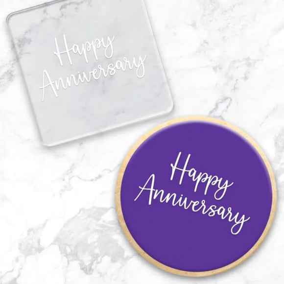 clear debosser with Happy Anniversary writing pattern beside a cookie with purple icing and white happy anniversary written displayed on a marble background