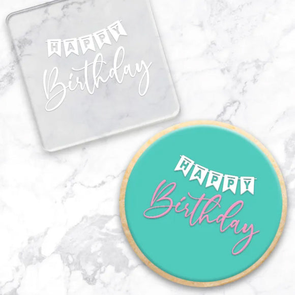 clear debosser with a Happy Birthday pattern beside a cookie with teal icing and happy birthday in pink and white  on a marble background