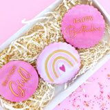 cookies with pink icing and rainbow pattern pained gold in a white gift box with ivory shredded paper and sprinkles scattered around