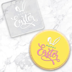 clear debosser with a happy easter with bunny ears pattern beside a cookie with yellow icing and pink, blue and white writing on a marble background