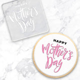 clear debosser with a Happy Mothers Day pattern beside a cookie with white icing and black and white writing on a marble background