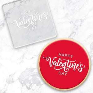 clear debosser with a happy Valentine's Day pattern beside a cookie with red icing and white writing on a marble background
