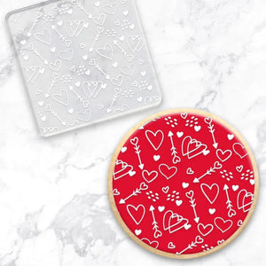 clear debosser with a heart valentine pattern beside a cookie with red icing and white writing on a marble background