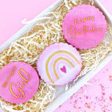 cookies with pink icing and rainbow pattern pained gold in a white gift box with ivory shredded paper and sprinkles scattered around