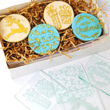 white box filled with brown shredded paper and cookies with Christmas patterns from debasers in gold, blue and ivory with debasers laid out below on a white background