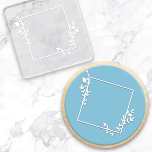 clear debosser with square frame pattern beside cookie with blue icing and white square pattern with a marble background